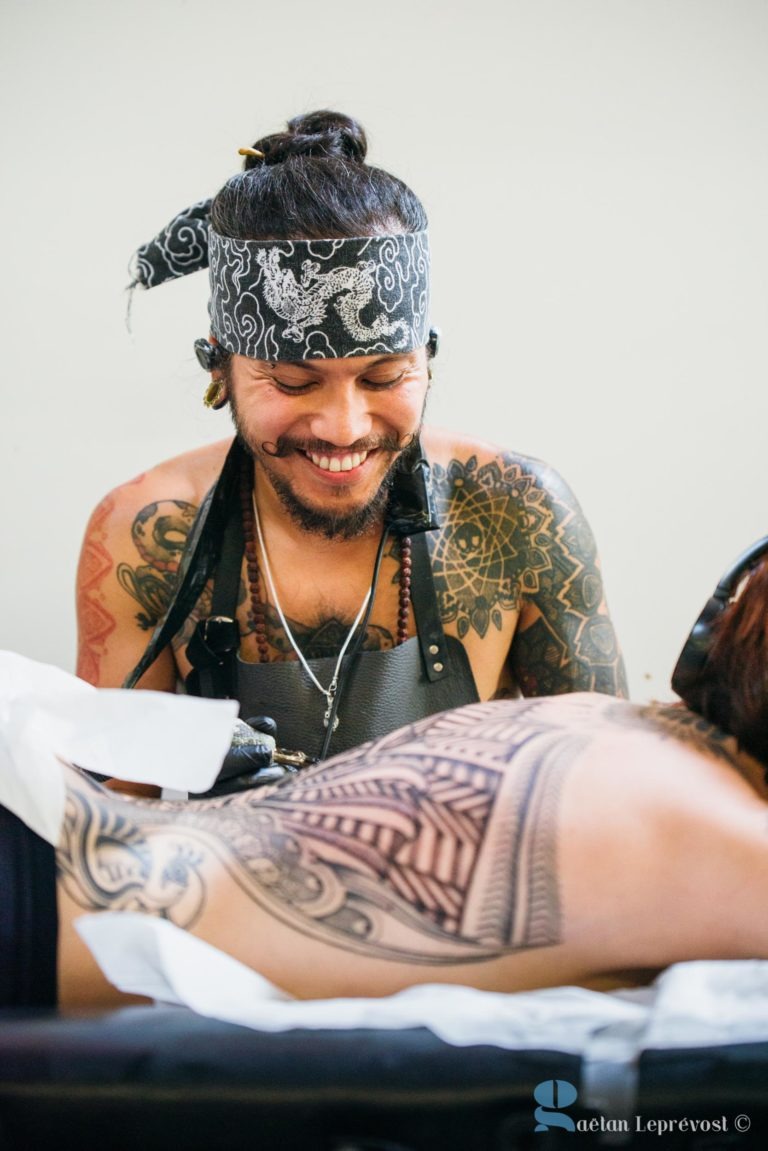 Un tatoueur avec un bandana à motifs sur la tête et des tatouages complexes sur les bras sourit tout en travaillant sur le dos d'un client au Salon Love Tattoo à La Teste-de-Buch. Le client est allongé sur le ventre, révélant un tatouage géométrique partiellement terminé sur le bas du dos. Le décor semble être un studio de tatouage.© Gaetan Leprévost.