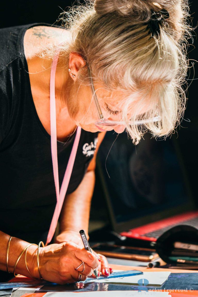 Une femme aux cheveux gris attachés en queue de cheval, portant des lunettes et une chemise noire, dessine attentivement sur une feuille de papier. Elle utilise un crayon et dispose de plusieurs marqueurs colorés et d'outils de dessin sur la table. L'arrière-plan est flou, laissant apparaître un ordinateur portable et une peinture colorée du Salon Love Tatoo La Teste-de-Buch.