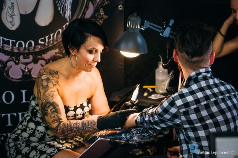 Un tatoueur aux cheveux courts et foncés et aux vêtements à motifs de crânes tatoue le bras d'une personne. Le bras gauche de l'artiste est fortement tatoué. Le client porte une chemise à carreaux. Une lampe éclaire le poste de travail du Salon Love Tattoo à La Teste-de-Buch. L'arrière-plan présente une affiche avec "Tattoo Shop" et des illustrations.