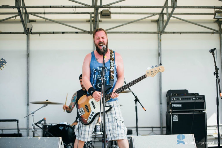 Un homme barbu, vêtu d'un débardeur bleu « Metal Heaven Old School » et d'un short à carreaux, chante et joue de la guitare basse avec passion sur une scène en plein air à La Teste-de-Buch. Derrière lui, un batteur joue, partiellement visible au milieu de l'échafaudage métallique. Des amplificateurs sont positionnés des deux côtés.