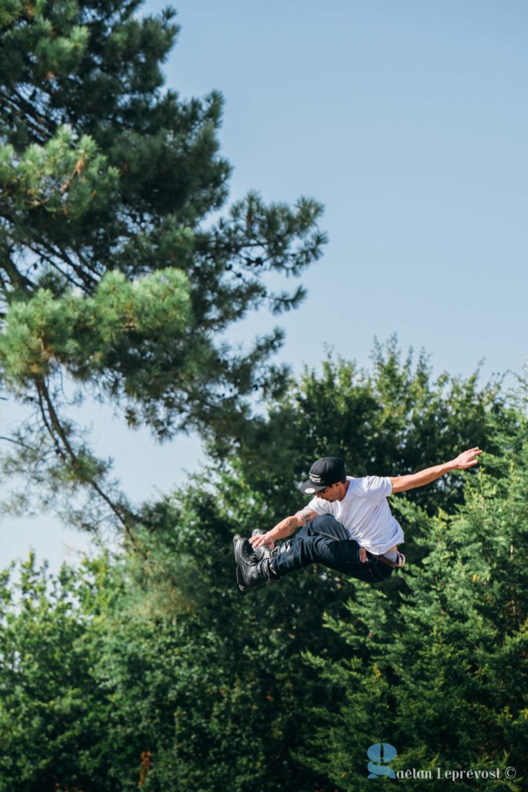 Un skateur est en plein vol et exécute une figure de haut vol. Il porte une chemise blanche, un pantalon noir et une casquette noire. En arrière-plan, il y a de grands arbres verts et un ciel bleu clair. La photo capture l'action et l'élévation du saut au-dessus du Salon Love & Tattoo à La Teste-de-Buch. Le filigrane du photographe se trouve dans le coin inférieur droit.