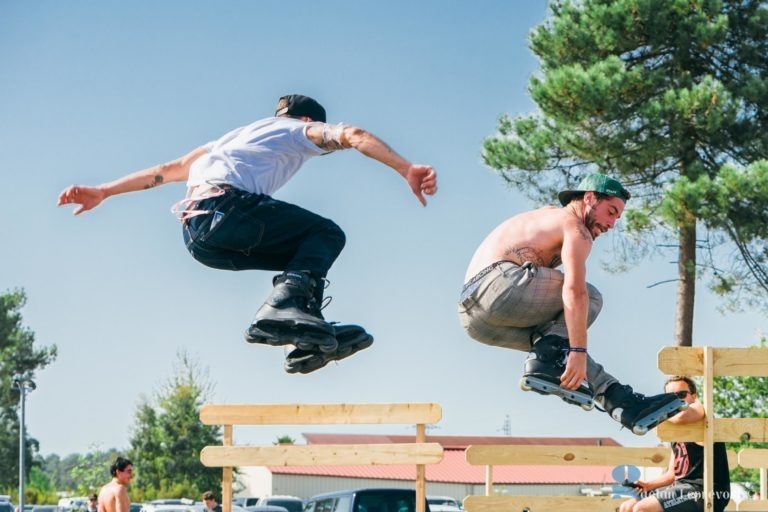 Deux hommes sont photographiés en plein vol alors qu'ils font du roller, exécutant des figures sur une structure en bois dans un parc extérieur près de La Teste-de-Buch. Tous deux portent des vêtements décontractés et un équipement de protection. La chemise d'un homme flotte alors qu'il saute. En arrière-plan, il y a des gens, des voitures, des arbres et un ciel bleu clair.