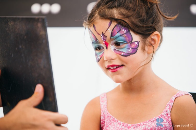 Une jeune fille au visage peint en papillon regarde son reflet dans un miroir à main. Le motif du papillon est coloré, avec des nuances de violet, de bleu et de rose, avec des points blancs et des étoiles en guise d'accents. Elle porte un débardeur à motifs roses et sourit doucement au Salon Love Tatoo à La Teste-de-Buch.