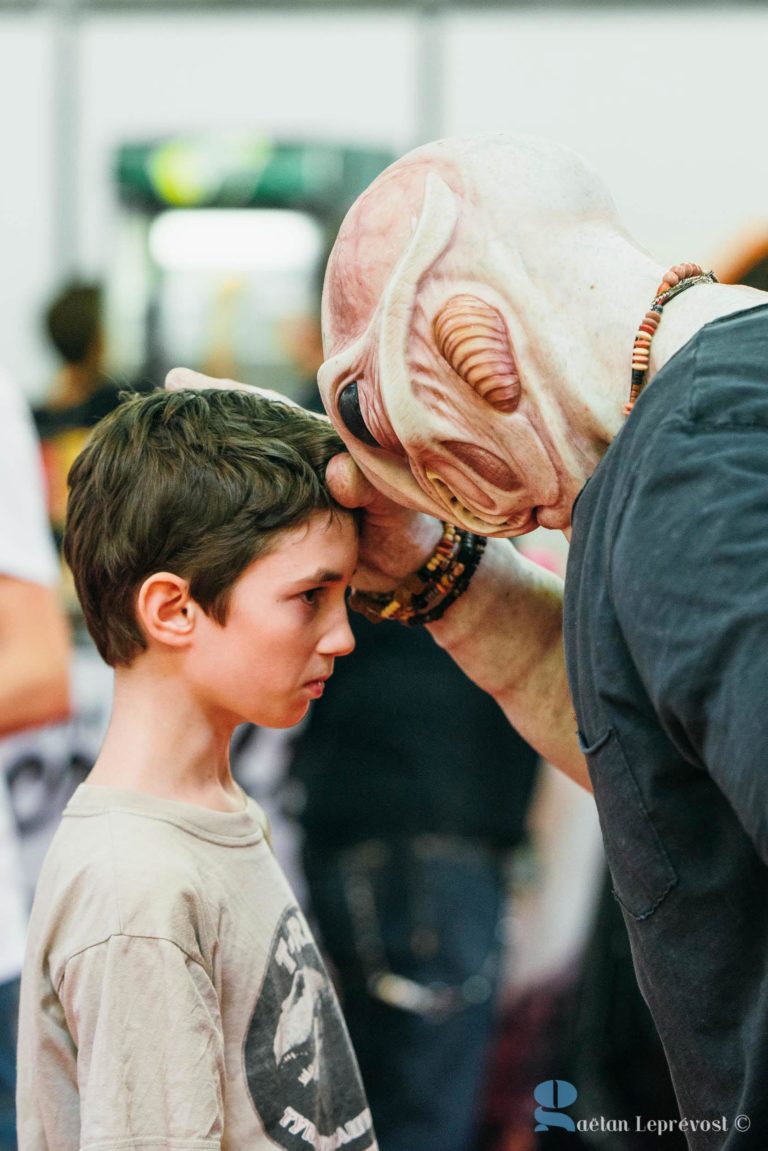 Un enfant, vêtu d'un t-shirt beige avec un imprimé dessus, interagit avec une personne portant un costume d'extraterrestre élaboré lors de l'événement Salon Love Tattoo à La Teste-de-Buch. L'extraterrestre, portant de grosses lunettes et des perles autour du cou, touche doucement le front de l'enfant avec le sien. Arrière-plan flou; texte : "© Gaetan LE PREVOST.