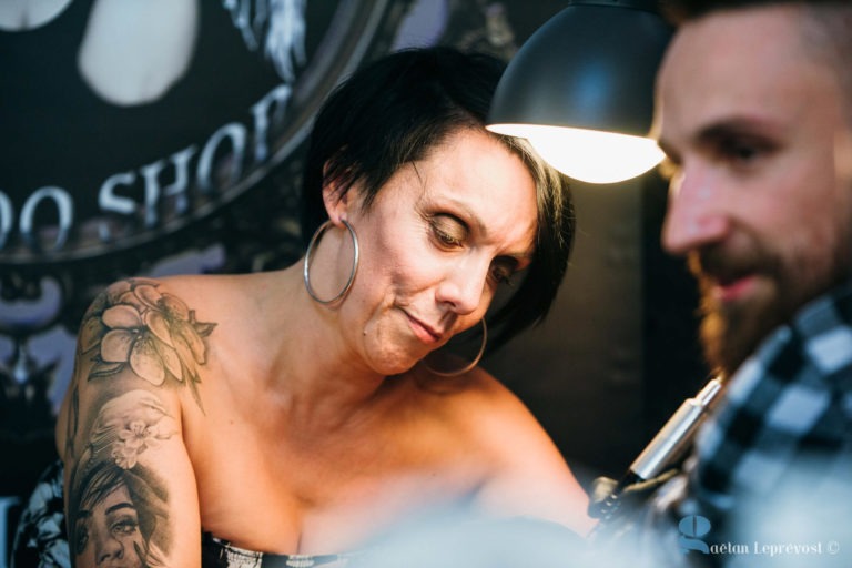 Une tatoueuse aux cheveux courts et foncés et aux grandes créoles se concentre sur le tatouage d'une cliente au Salon Love Tattoo de La Teste-de-Buch. Elle a un tatouage sur le bras représentant un visage. La scène est faiblement éclairée par une lampe au-dessus d'elle, et l'arrière-plan montre un logo de salon de tatouage partiellement visible. La cliente est légèrement floue.