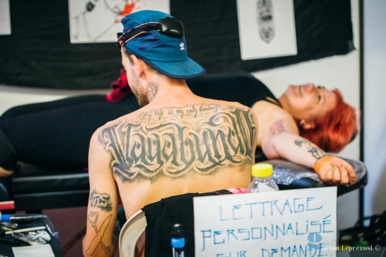 Un tatoueur, coiffé d'un chapeau à l'envers, est vu de dos, dévoilant un grand tatouage sur le haut de son dos sur lequel est écrit "Vagabond". Il tatoue une personne aux cheveux roux allongée sur une table au Salon Love Tatoo La Teste-de-Buch. Une pancarte au premier plan indique "LETTTRAGE PERSONNALISÉ SUR DEMAND". Des motifs de tatouage sont visibles sur le