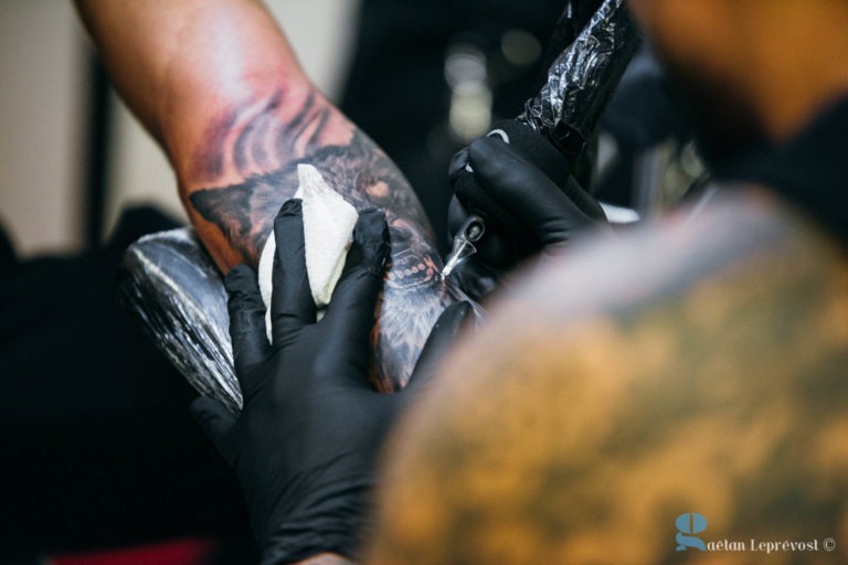 Un artiste aux bras tatoués, portant des gants noirs, utilise une machine à tatouer pour tatouer l'avant-bras d'une autre personne au Salon Love Tattoo à La Teste-de-Buch. Le dessin semble être un portrait. La personne qui se fait tatouer a son bras posé sur un support rembourré. L'arrière-plan est flou, attirant l'attention sur le tatouage en cours.