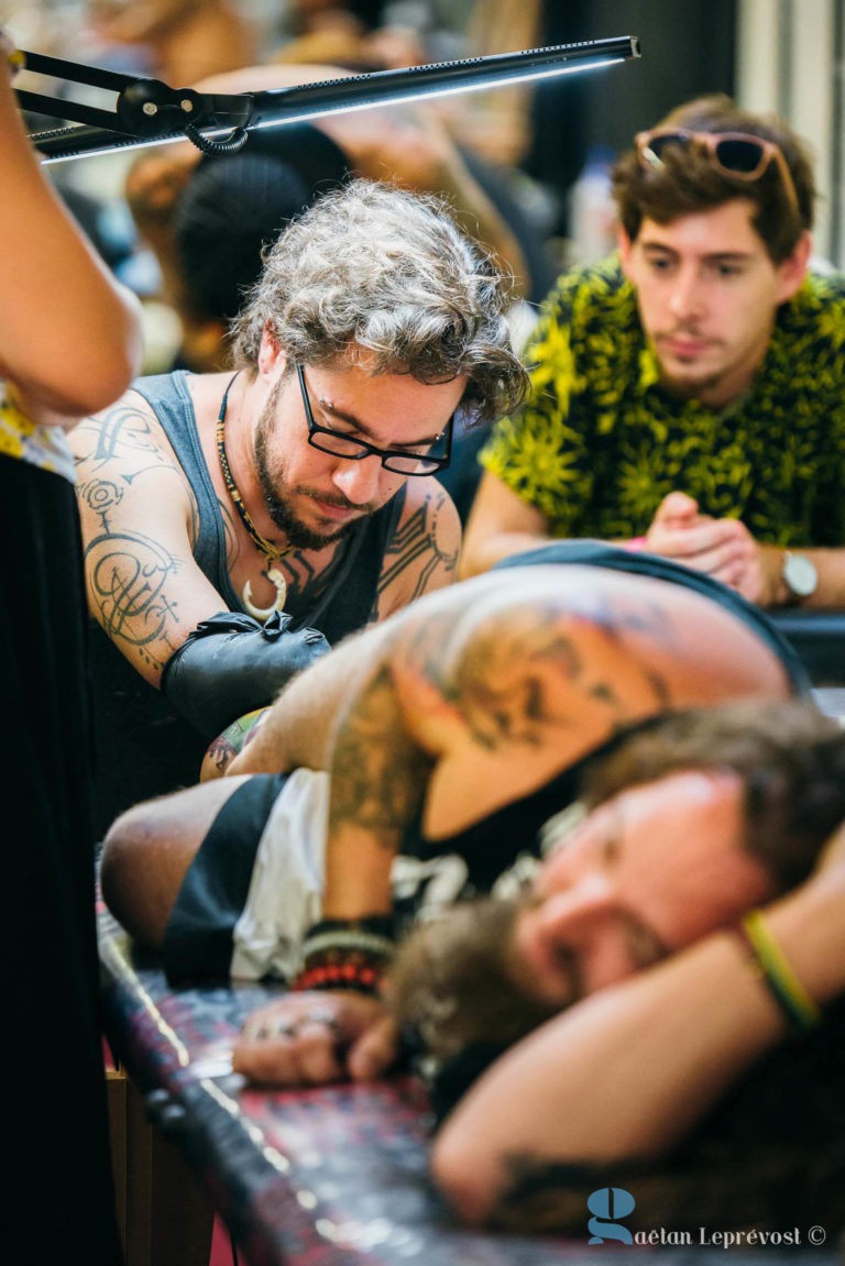 Un homme portant des lunettes et des tatouages sur les bras se concentre sur le tatouage d'une autre personne allongée sur une table du Salon Love Tattoo à La Teste-de-Buch. Un autre homme portant une moustache et une chemise à motifs jaunes observe attentivement depuis l'arrière-plan. Du matériel de tatouage et une lampe sont visibles dans l'espace de travail.