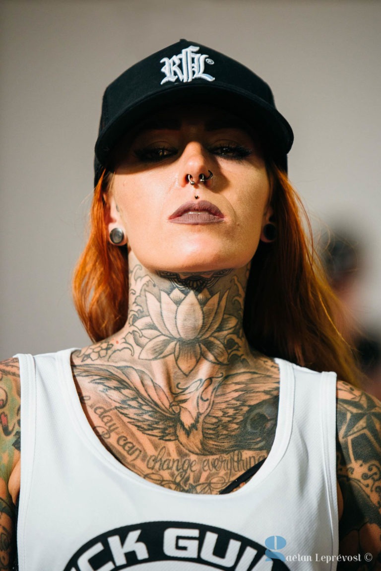 Une personne aux longs cheveux roux et aux tatouages étendus du Salon Love Tattoo La Teste-de-Buch porte une casquette noire avec des lettres blanches et un débardeur blanc. Elle a plusieurs piercings, dont un anneau de septum et des boucles d'oreilles de gros calibre. L'arrière-plan de l'image est flou. Crédit photo : Gaétan Leprevost.