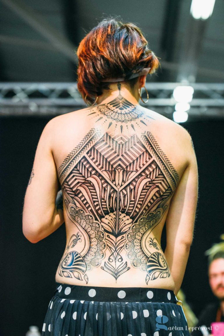 Une femme aux cheveux bruns courts arbore un tatouage élaboré dans le dos avec des motifs géométriques complexes et symétriques. Son dos nu met en valeur le dessin détaillé à l'encre noire, s'étendant de son cou jusqu'au bas de son dos. Elle porte une jupe à pois noirs et blancs. Le fond sombre crée un cadre parfait au Salon Love Tattoo La Teste-de-Buch.