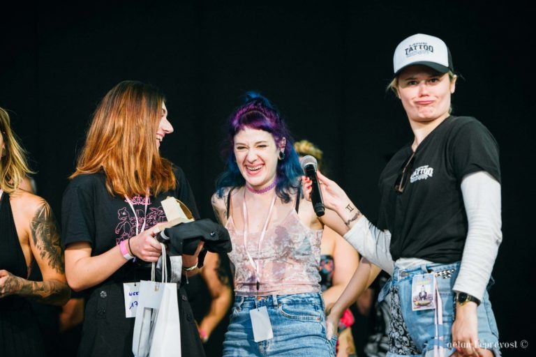 Trois femmes sont sur scène au Salon Love, souriantes et riantes. La femme de gauche a les cheveux bruns et tient une chemise noire et un sac cadeau. La femme au centre a les cheveux violets et bleus avec des tatouages sur les bras. La femme de droite tient un micro, porte une casquette et une chemise noire avec le mot "TATTOO".