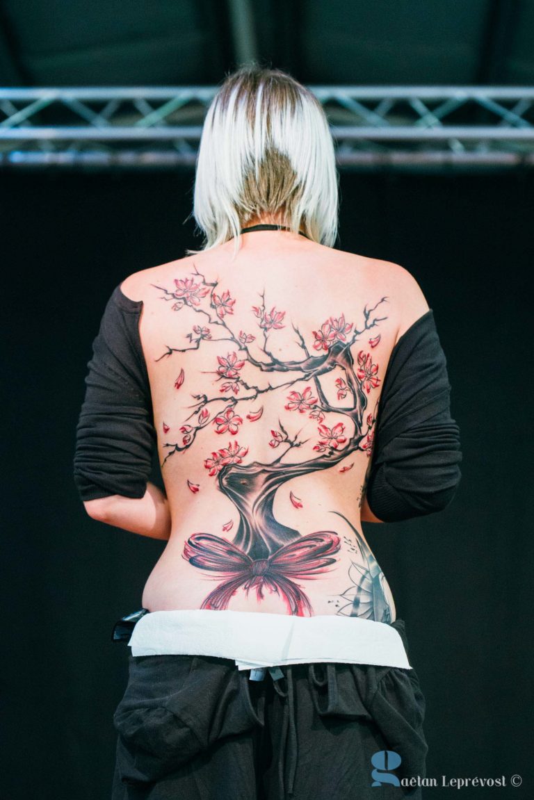 Une personne aux cheveux blonds platine courts est représentée de dos, affichant fièrement un grand tatouage complexe d'un cerisier en fleurs créé au Salon Love Tattoo La Teste-de-Buch. Le tatouage s'étend du bas du dos jusqu'au haut des épaules et représente une balançoire suspendue à une branche. La personne porte un haut noir et un pantalon blanc.