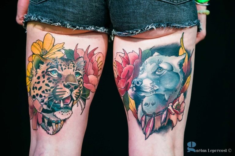 Une paire de cuisses tatouées avec des motifs animaliers distincts, réalisées au Salon Love Tattoo à La Teste-de-Buch. La cuisse gauche présente un léopard grognant entouré de fleurs jaunes et roses. La cuisse droite affiche un loup calme encadré de fleurs rouges et jaunes. Les deux tatouages sont colorés et détaillés, la personne portant un short noir.