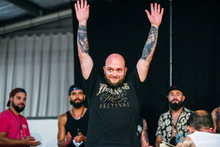 Un homme barbu se tient debout, les bras levés, souriant. Il a des tatouages sur les deux bras et porte un t-shirt noir avec des lettres dorées sur lesquelles est écrit « Festival de la barbe et de la moustache ». Plusieurs autres hommes barbus et moustachus sont visibles en arrière-plan, certains applaudissant. Le décor semble être le Salon Love Tatoo à La Teste-de-Buch.