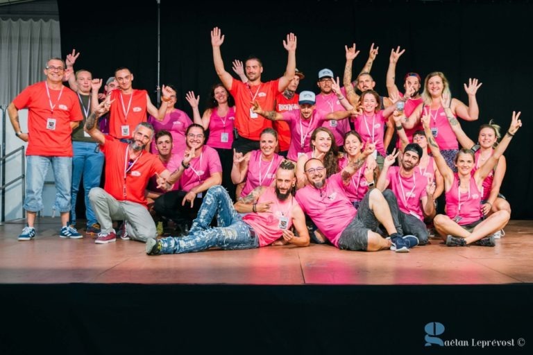 Un groupe de personnes pose sur une scène, la plupart portant des chemises roses et quelques-unes des chemises rouges, toutes ornées de badges nominatifs. Elles sourient, font des signes de paix avec leurs mains et dégagent une atmosphère amicale et joyeuse. Une personne est allongée sur le sol de manière enjouée. Le fond est noir, symbolisant le Salon Love Tatoo de La Teste-de-Buch.