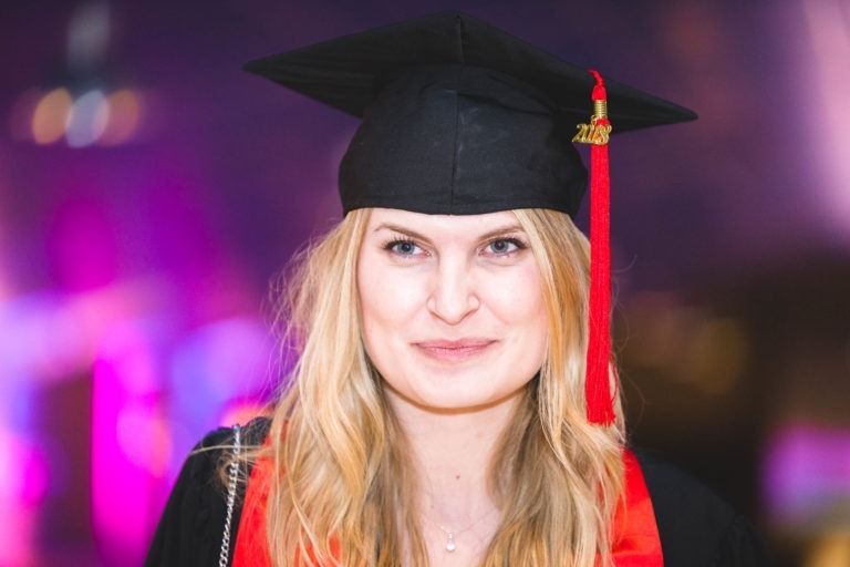 Une jeune femme aux longs cheveux blonds sourit à la caméra tout en portant un bonnet de remise de diplôme noir avec un pompon rouge et or, et une toge noire avec une étole rouge. L'arrière-plan est flouté par des lumières violettes et jaunes, suggérant l'ambiance festive de la Soirée de remise des diplômes EBBS Bordeaux.