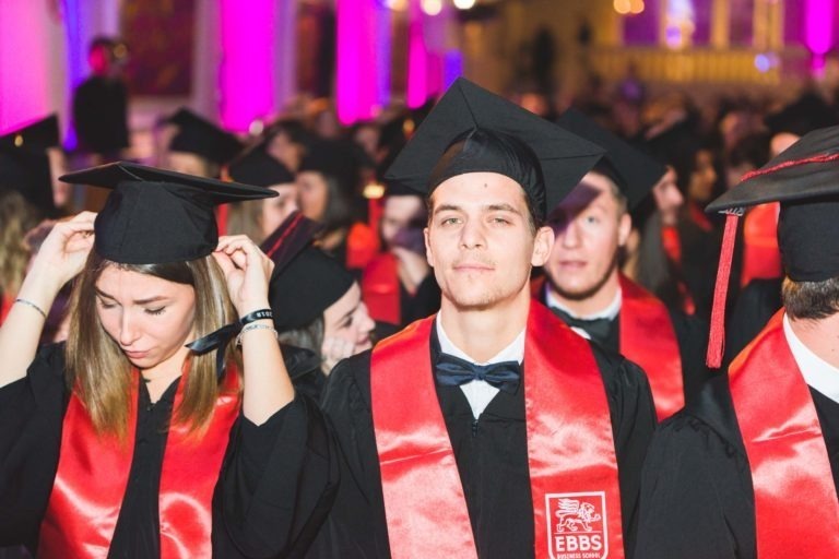 Un groupe de diplômés vêtus de toges noires et de mortiers avec des étoles rouges, ornés d'un emblème sur lequel est inscrit « EBBS », célèbrent la remise des diplômes EBBS Bordeaux. L'accent est mis sur un jeune homme au centre, légèrement souriant, et une jeune femme ajustant sa casquette. L'arrière-plan est rempli d'autres diplômés et illuminé de manière festive dans des tons violets.