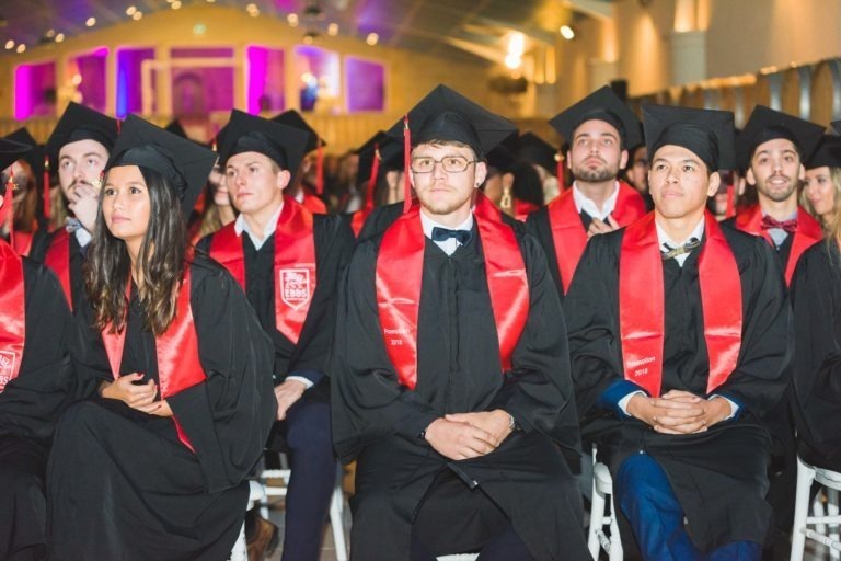 Un groupe d'étudiants portant des toges noires et des casquettes avec des étoles rouges et blanches sont assis en rangées lors de la Soirée de remise des diplômes EBBS Bordeaux. Ils semblent attentifs et concentrés, avec une scène et des lumières décoratives en arrière-plan. La disposition des sièges se compose de chaises blanches placées dans un hall spacieux.