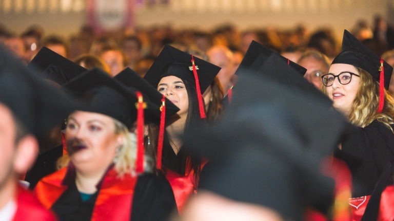 Un grand groupe de diplômés portant des toges et des casquettes noires avec des glands et des écharpes rouges sont assis pendant la cérémonie de remise des diplômes de l'EBBS Bordeaux. L'arrière-plan est rempli de personnes, probablement des membres de la famille et des amis, flous. L'ambiance semble festive et concentrée.