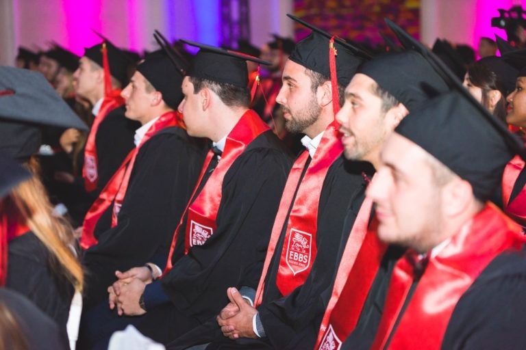 Un groupe de diplômés portant des toges et des casquettes noires avec des écharpes rouges sont assis en rangées. Ils regardent attentivement vers l'avant lors de la cérémonie de remise des diplômes EBBS Bordeaux. L'ambiance est festive avec des lumières colorées en arrière-plan. Les écharpes rouges affichent un logo et l'acronyme « EBBS ».