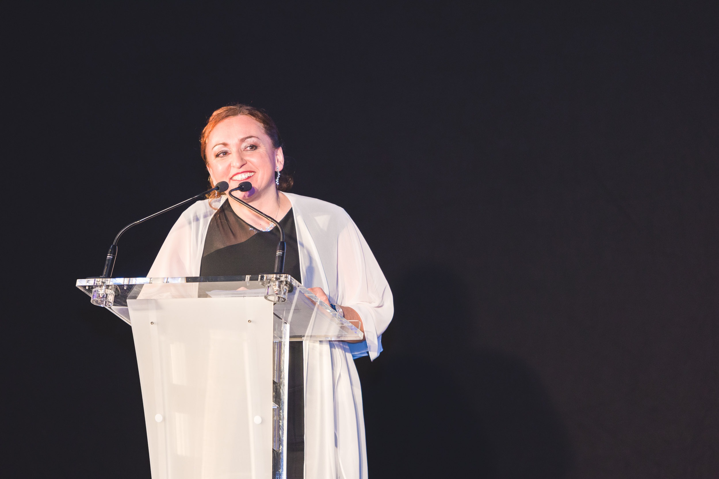 Une femme aux cheveux châtain clair tirés en arrière, portant un châle blanc sur une robe noire, se tient debout sur un podium en acrylique transparent et parle dans un micro. Elle sourit et semble prononcer un discours lors de la Soirée de remise des diplômes EBBS Bordeaux sur un fond sombre.