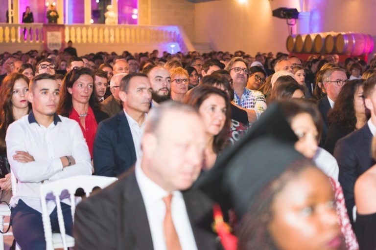 Un grand groupe de personnes est assis les uns à côté des autres dans un auditorium intérieur pour la soirée de remise des diplômes de l'EBBS Bordeaux. Le public est diversifié, avec des individus d'âges et de tenues différents. Au premier plan, certains visages sont flous en raison de la profondeur de champ. L'arrière-plan présente un balcon avec des personnes debout et un éclairage lilas doux.