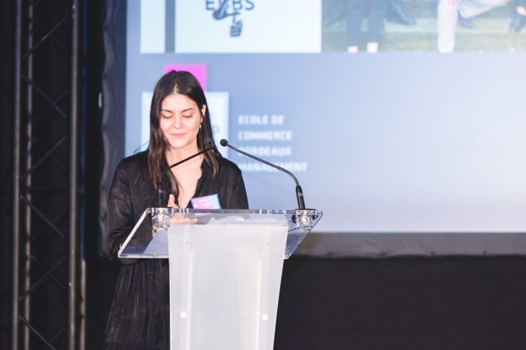 Une femme se tient debout sur un podium dégagé et parle dans un micro. Elle a de longs cheveux noirs et est vêtue d'une tenue noire. Derrière elle, un écran de projection diffuse une image et un texte, bien que le contenu exact ne soit pas clair. Le décor semble être la Soirée de remise des diplômes de l'EBBS Bordeaux.