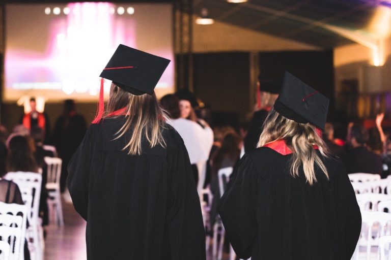 Deux diplômés en toge noire et casquette à pompons rouges se tiennent face à la scène lors de la Soirée de remise des diplômes EBBS Bordeaux. Ils se trouvent dans une grande salle remplie de chaises blanches et de participants. La scène en arrière-plan montre un écran avec des lumières colorées, capturant une atmosphère festive et formelle.