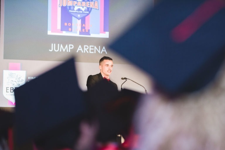 Un homme en costume noir s'exprime sur une estrade devant un grand écran affichant « JUMP ARENA » et « BOOST YOUR BOUNCE » avec un graphisme de trampoline. Les toques de diplômés portées par les membres du public indiquent qu'il s'agit de la soirée de remise des diplômes de l'EBBS Bordeaux. Un logo EBE est partiellement visible sur l'écran.