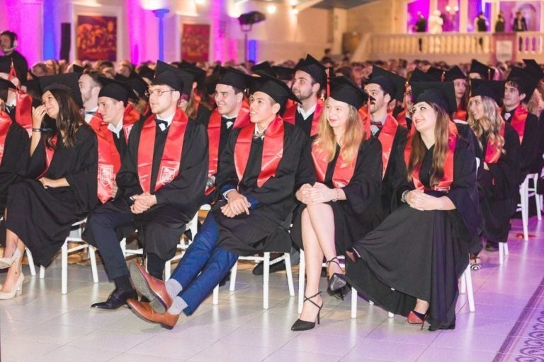 Un groupe d'étudiants portant des toges noires, des mortiers et des écharpes rouges sont assis en rangées lors de la Soirée de remise des diplômes EBBS Bordeaux. Ils se trouvent dans une salle bien éclairée avec des éclairages violets. Le public, flou en arrière-plan, semble observer l'événement avec attention.