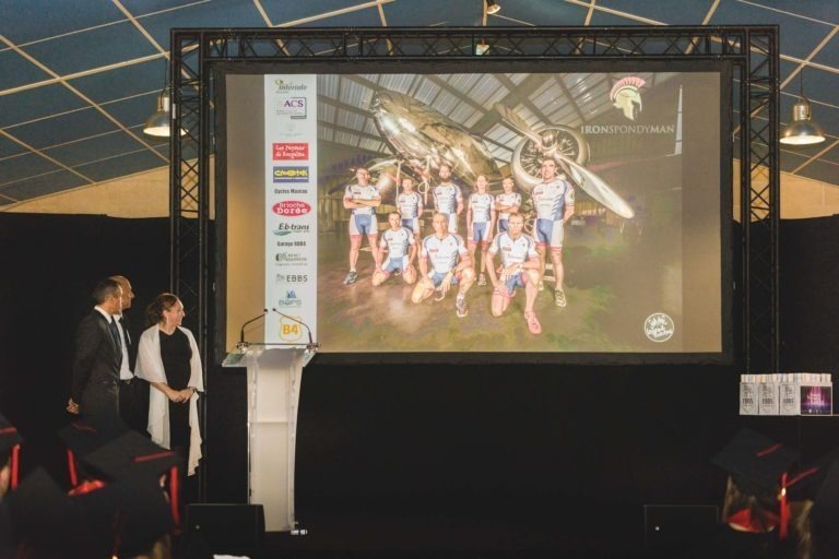 Un homme et une femme se tiennent debout sur une estrade et s'adressent à un public. Derrière eux, un grand écran diffuse l'image d'une équipe sportive posant avec des vélos. Les logos des sponsors sont affichés sur un côté de l'écran. La salle, qui accueille la Soirée de remise des diplômes EBBS Bordeaux, est dotée d'un haut plafond avec des lumières de scène vives au-dessus.