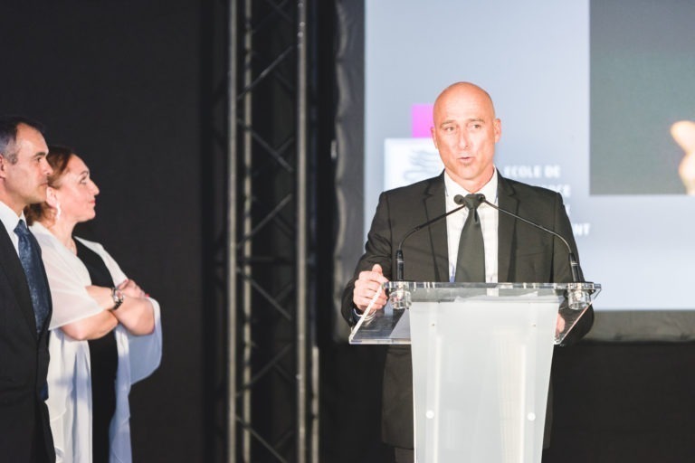 Un homme chauve en costume noir et chemise blanche parle dans un micro sur un podium dégagé. Il est sur scène, avec un écran diffusant une partie d'une image et d'un texte derrière lui. Trois personnes se tiennent à sa gauche, l'écoutent attentivement, avec une femme croisant les bras lors de la Soirée de remise des diplômes EBBS Bordeaux.