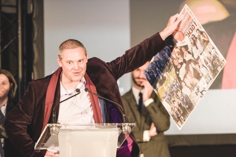 Un homme vêtu d'un blazer sombre se tient debout sur un podium dégagé, brandissant une grande affiche avec des images et du texte. Il s'adresse à un public, avec une présentation projetée en arrière-plan. Une autre personne, partiellement cachée et en costume, se trouve à l'arrière-plan. Le décor semble être la Soirée de remise des diplômes de l'EBBS Bordeaux.