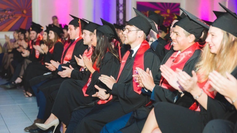 Une rangée de diplômés universitaires portant des toges et des casquettes noires avec des étoles rouges applaudissent à la Soirée de remise des diplômes EBBS Bordeaux. Ils se déroulent à l'intérieur, avec un éclairage coloré et des sièges bondés visibles en arrière-plan. Le groupe comprend à la fois des hommes et des femmes, tous semblant heureux et festifs.