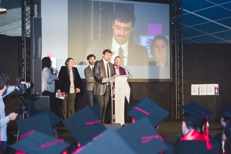 Un homme en costume s'exprime sur une estrade, à côté de trois personnes. Derrière eux, un grand écran diffuse un gros plan de l'orateur. Le public, coiffé de toques de diplômés, regarde attentivement. La soirée de remise des diplômes EBBS Bordeaux semble formelle, mettant l'accent sur l'orateur et les diplômés.