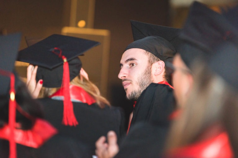 Un groupe de diplômés vêtus de toges et de casquettes noires avec des pompons et des doublures rouges sont assis, assistant à la Soirée de remise des diplômes EBBS Bordeaux. Un diplômé, un homme barbu en gros plan, regarde vers sa droite. L'arrière-plan est flou, soulignant l'atmosphère formelle et festive qui entoure les diplômés.