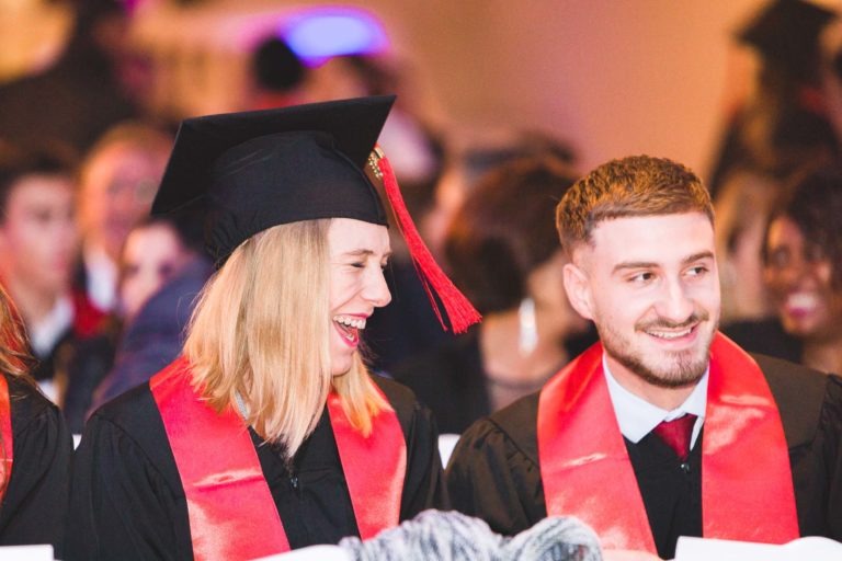 Deux diplômés portant des toges et des casquettes noires avec des écharpes rouges sourient assis ensemble lors de la Soirée de remise des diplômes de l'EBBS Bordeaux. La personne à gauche a les cheveux blonds jusqu'aux épaules et le rire est visible sur son visage. La personne à droite a les cheveux bruns courts et une barbe taillée, souriant chaleureusement. Le public est flou en arrière-plan.