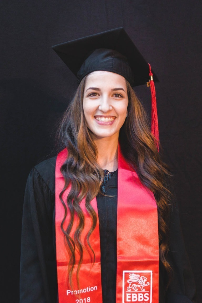 Une personne souriante en tenue de remise de diplôme se tient devant un fond uni. Elle porte une toque noire avec un gland rouge et une étole rouge sur laquelle sont inscrits en blanc les mots « EBBS » et « Promotion 2018 », ainsi qu'un logo représentant un lion. La personne, aux cheveux longs et ondulés, a assisté à la Soirée de remise des diplômes EBBS Bordeaux.