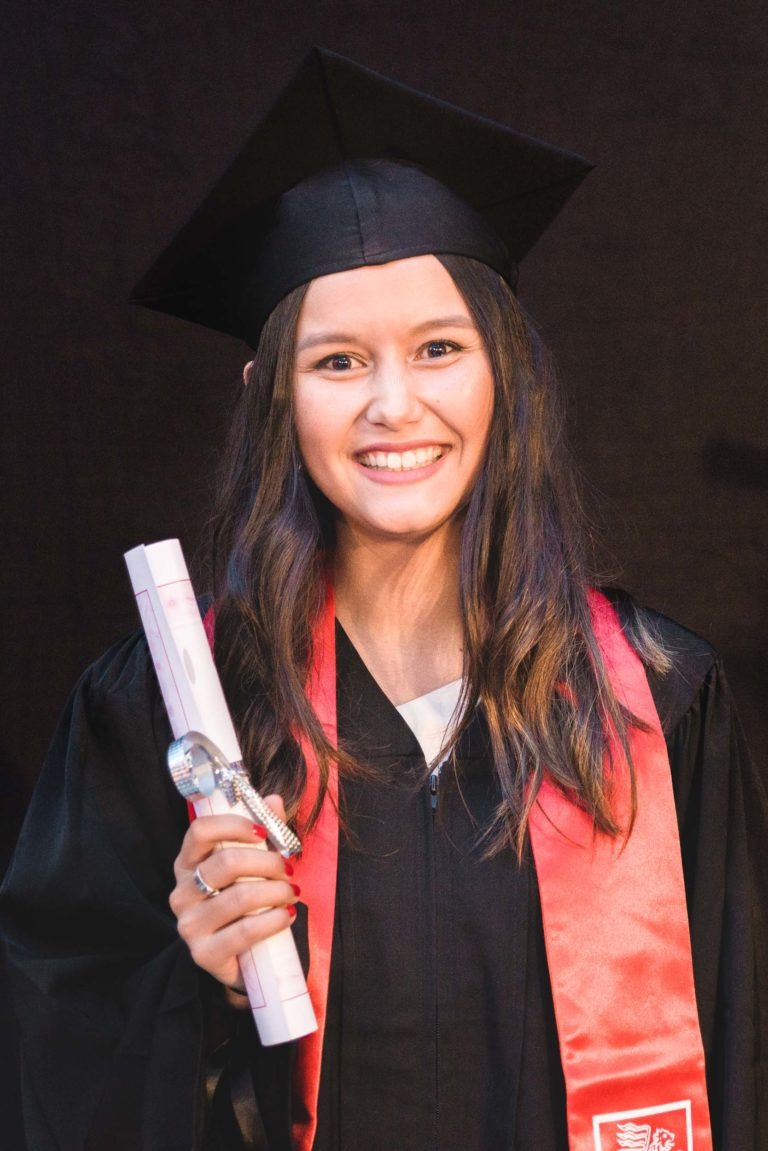 Une jeune femme aux cheveux longs et foncés se tient debout, souriante, dans sa tenue de remise des diplômes lors de la soirée de remise des diplômes de l'EBBS Bordeaux. Elle porte une toge et un bonnet noirs avec une étole rouge et tient un diplôme roulé dans sa main droite. L'arrière-plan est uni et sombre, faisant d'elle le point central de l'image.