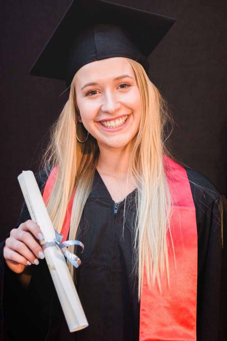 Une jeune femme aux longs cheveux blonds sourit et porte une toge et un bonnet noirs avec une étole rouge. Elle tient dans sa main droite un diplôme enroulé et noué avec un ruban, célébrant la Soirée de remise des diplômes EBBS Bordeaux. L'arrière-plan est sombre, mettant en valeur son visage et sa tenue lumineuse.