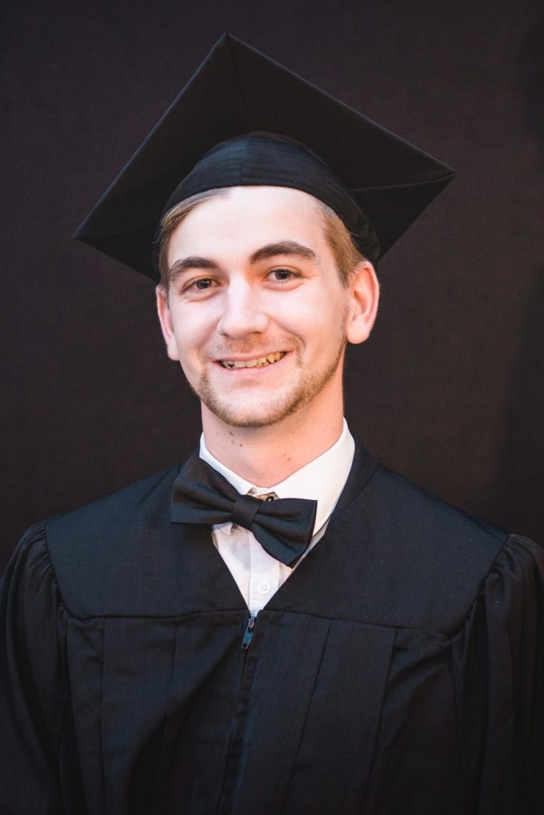 Un jeune homme portant une toge et un chapeau de remise de diplômes sourit à la caméra. Il porte un chapeau noir, une robe noire et un nœud papillon noir sur une chemise blanche. L'arrière-plan est uni et sombre, évoquant l'esprit de fête de la Soirée de remise des diplômes EBBS Bordeaux.