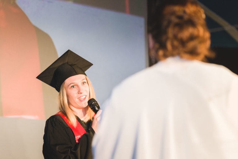 Une femme en tenue de remise de diplôme, comprenant une toge et un bonnet noirs avec une étole rouge, parle dans un micro lors de la soirée de remise des diplômes de l'EBBS Bordeaux. Elle fait face à une personne en toge blanche, qui apparaît floue au premier plan. Un grand écran diffusant une partie de la scène est visible derrière eux.