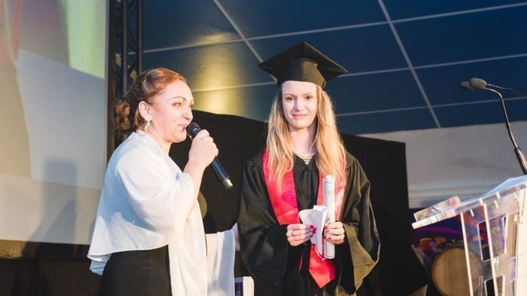 Une femme vêtue d'un châle blanc parle dans un micro sur une scène, en faisant des gestes de la main. À côté d'elle, une jeune femme portant une toge et un bonnet noirs et une étole rouge tient un diplôme enroulé. Elles se tiennent sous le dais bleu de la Soirée de remise des diplômes EBBS Bordeaux, éclairées par les lumières de la scène.