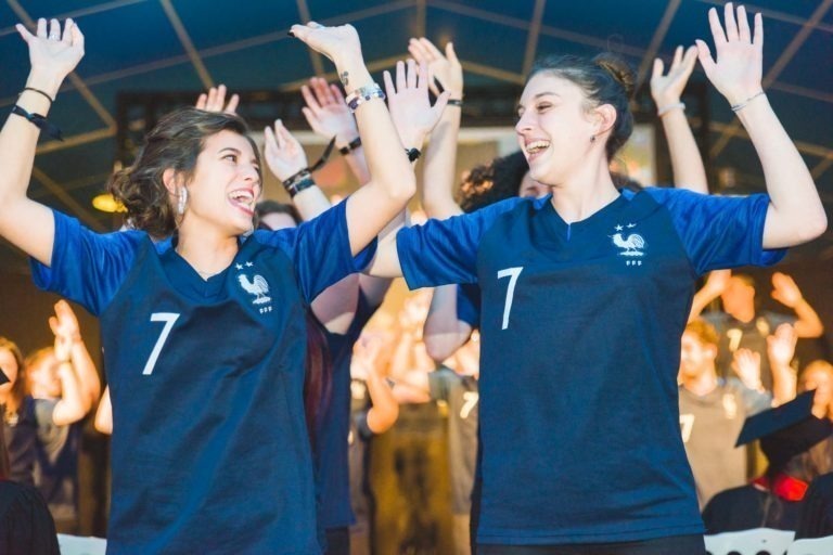 Deux femmes, toutes deux aux cheveux bruns attachés, sourient et lèvent les mains en signe de célébration lors de la soirée de remise des diplômes de l'EBBS Bordeaux. Elles portent des maillots bleu foncé assortis avec le numéro 7 blanc dessus. Derrière elles, un groupe de personnes portant des maillots sombres lèvent également les mains à l'unisson. L'arrière-plan est un intérieur.