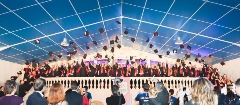 Un grand groupe de diplômés en toge noire avec ceinture rouge célèbre la Soirée de remise des diplômes EBBS Bordeaux en jetant leurs toques en l'air. Ils se tiennent sur une scène avec une balustrade blanche tandis qu'un public en contrebas, certains tenant des appareils photo, capture l'instant. Un plafond bleu avec un motif en grille est au-dessus.