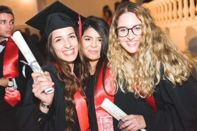 Trois diplômés sourient à la caméra, vêtus de toges noires et d'étoles rouges lors de la soirée de remise des diplômes de l'EBBS Bordeaux. La femme à gauche tient un diplôme, celle du milieu se tient près d'elle et celle à droite a les cheveux blonds bouclés et des lunettes, tenant également un diplôme. D'autres personnes sont visibles à l'arrière-plan.