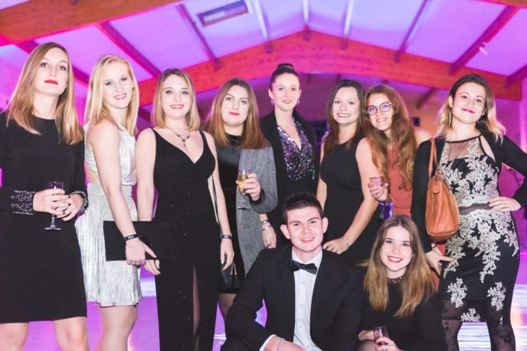 Un groupe de onze jeunes adultes, hommes et femmes, habillés en tenue de soirée, posent pour une photo lors d'une soirée de remise des diplômes de l'EBBS Bordeaux. Les femmes portent des robes élégantes de différents styles et couleurs, tandis que les hommes sont en costume et nœud papillon. L'arrière-plan présente un haut plafond avec des poutres en bois et un éclairage violet.