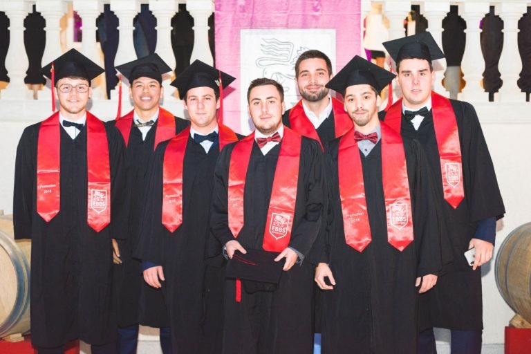 Sept diplômés, vêtus de toges noires et d'écharpes rouges, se tiennent en rang, souriant à la caméra lors de la soirée de remise des diplômes de l'EBBS Bordeaux. Quatre d'entre eux tiennent leurs diplômes devant une balustrade blanche et un fond rose. Ils portent tous des toges noires, célébrant leur réussite.