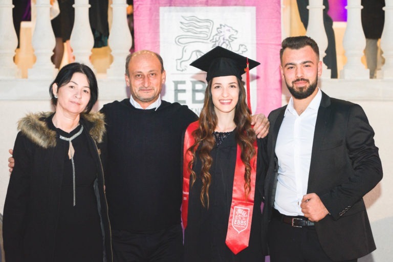 Une famille de quatre personnes se tient devant une banderole « EBBS ». Le diplômé, coiffé d'une toge et d'un bonnet noirs avec une ceinture rouge, se tient entre deux hommes en costume et une femme en veste noire bordée de fourrure. Tous sourient et regardent la caméra, célébrant la soirée de remise des diplômes EBBS Bordeaux.