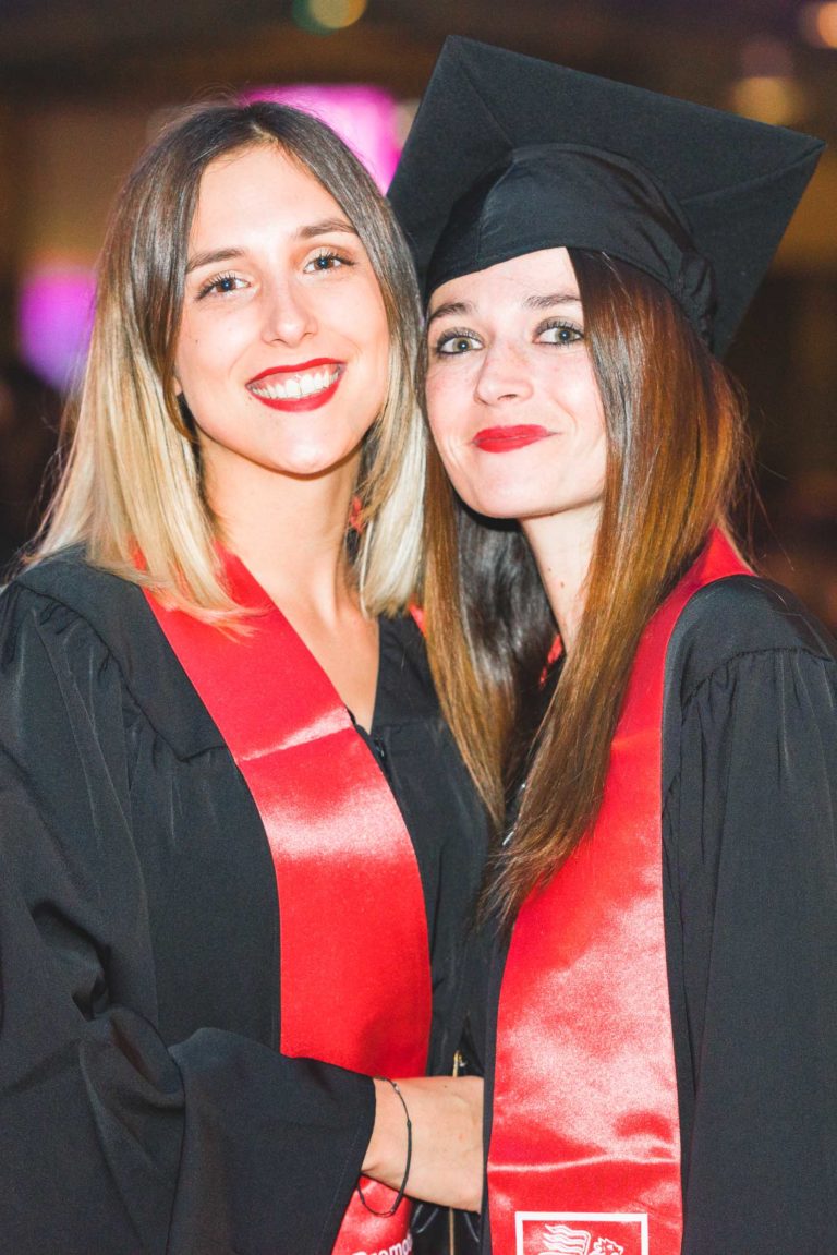 Deux femmes sourient à la caméra, vêtues de toges noires avec des ceintures rouges. La femme de gauche a les cheveux blonds jusqu'aux épaules, et celle de droite a les cheveux bruns longs et raides et porte une toque de diplômé. Elles se tiennent côte à côte, bras dessus, bras dessous, lors de la Soirée de remise des diplômes de l'EBBS Bordeaux.